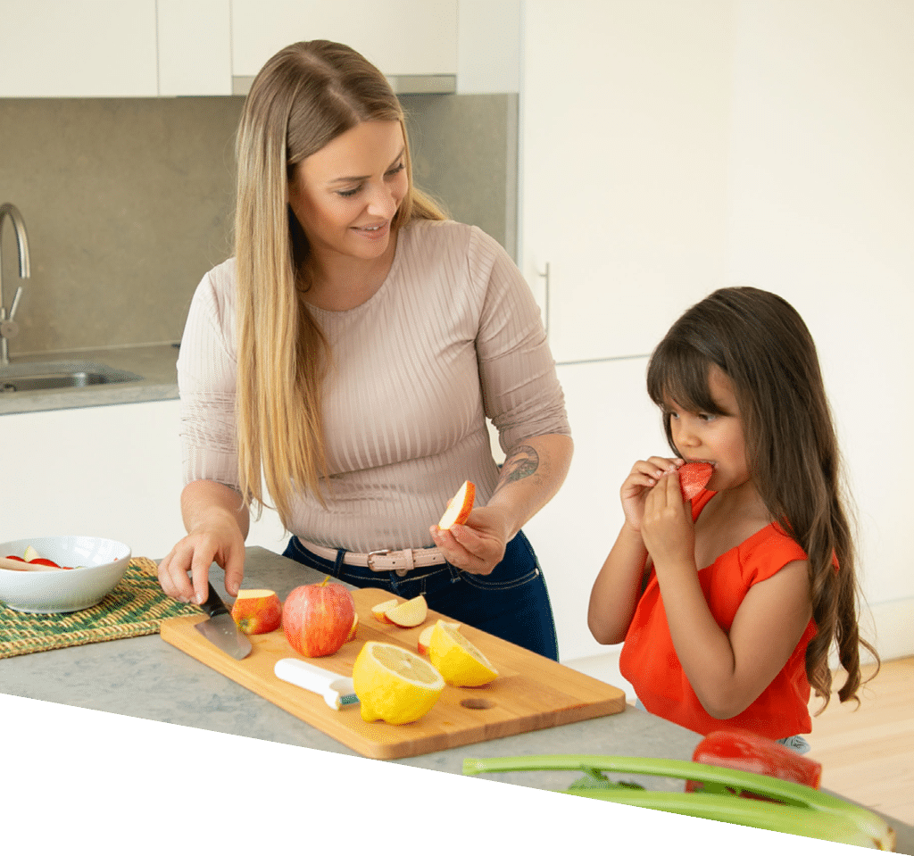 Mom giving daughter to taste apple slice while cooking salad  Girl and her mother cooking together, cutting fresh fruits and vegs on chopping board in kitchen  Family cooking concept