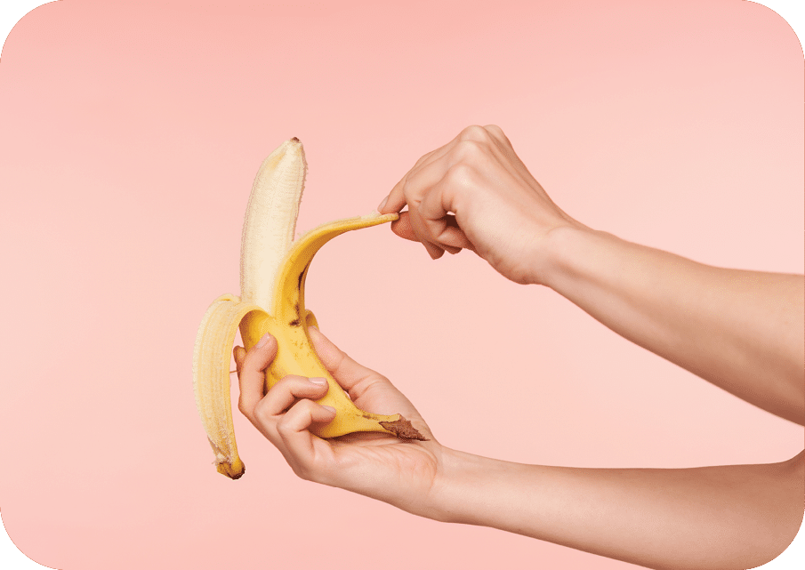 Studio shot of elegant woman's hands holding banana while peeling it and going to bite, having healthy breakfast while being isolated over pink background