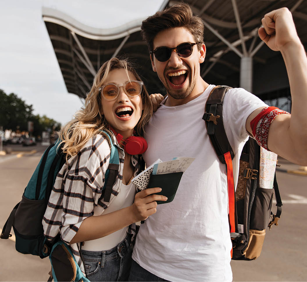 Attractive blonde woman in sunglasses and man in white tee smiles and takes selfie near airport. Portrait of happy travelers with backpacks.