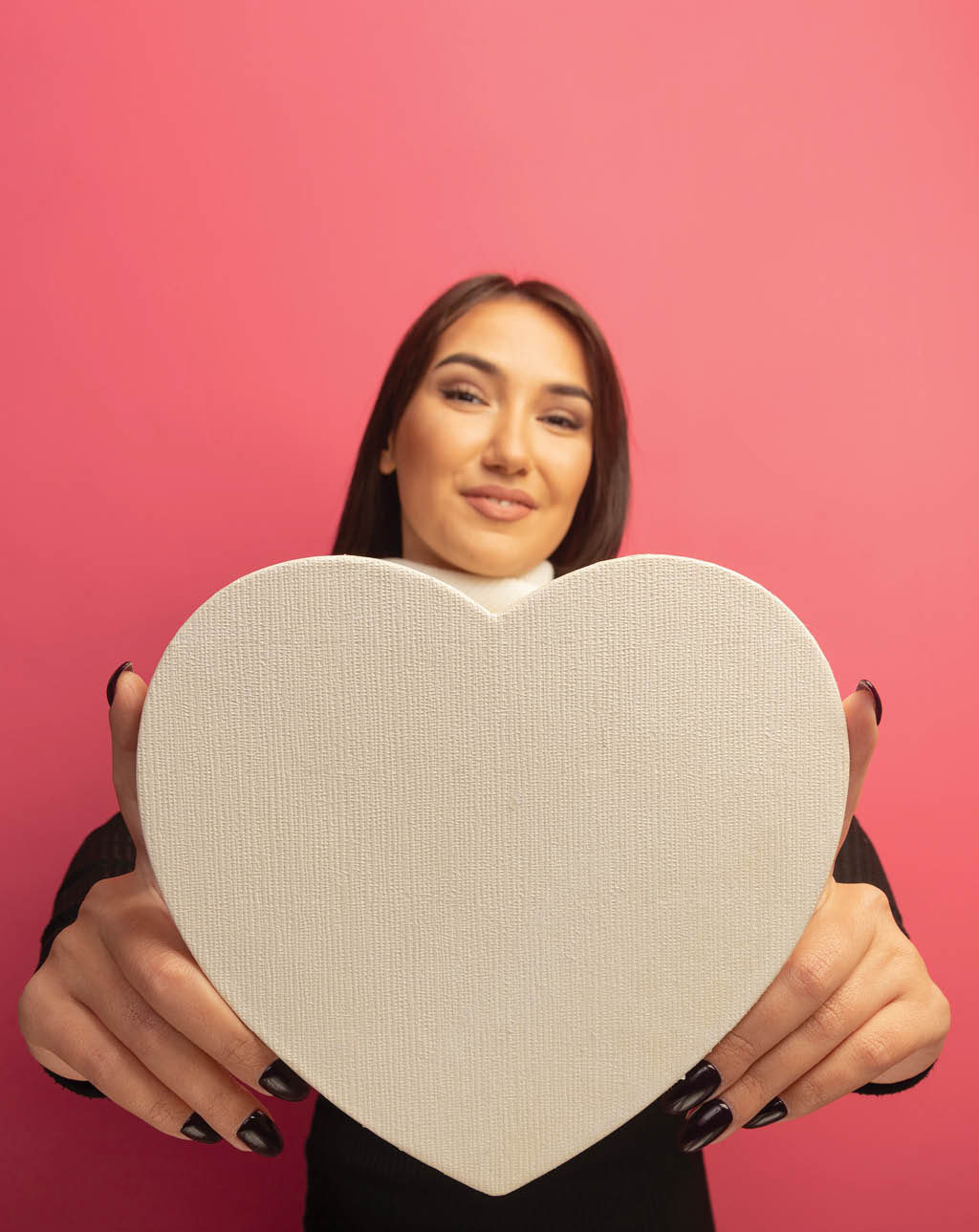 young woman with white scarf showing cardboard heart looking at camera smiling cherfully standing over pink background