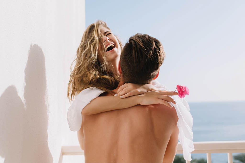 Laughing girl in white blouse having fun on terrace spending summer vacation with her boyfriend  Beautiful happy couple hugging on balcony with sea views on honeymoon on exotic resort