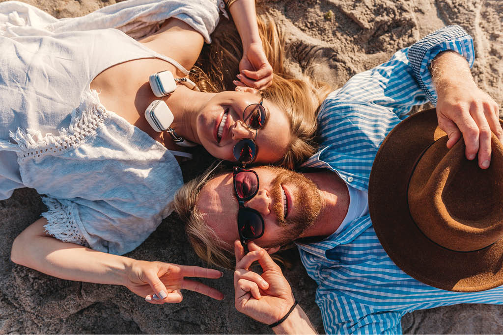 view from above on young attractive smiling happy man and woman in sunglasses lying on sand beach, romantic couple by the sea on sunset, boho hipster style outfit, friends having fun together