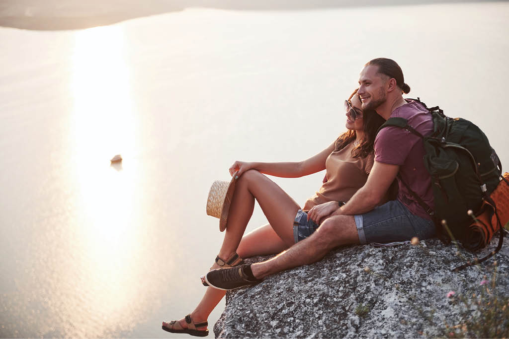 Hugging couple with backpack sitting on top of rock mountain enjoying view coast a river or lake  Traveling mountains and coast, freedom and active lifestyle concept 