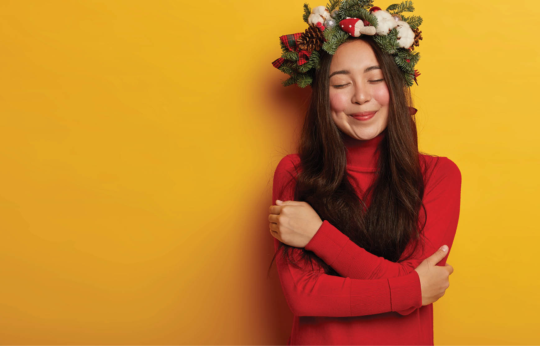 Tender woman with long dark hair hugs herself, enjoys comfort in new jumper, keeps eyes closed from pleasant emotions, smiles tenderly, isolated over yellow wall, copy space for your promotion