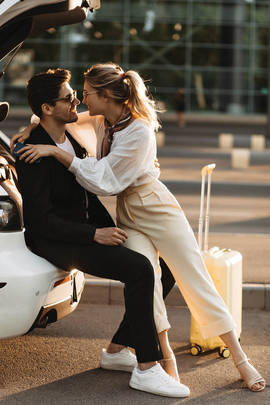 Pretty woman in white outfit smiles widely and hugs boyfriend  Brunette man in black suit looks at girlfriend and sits in car near airport 