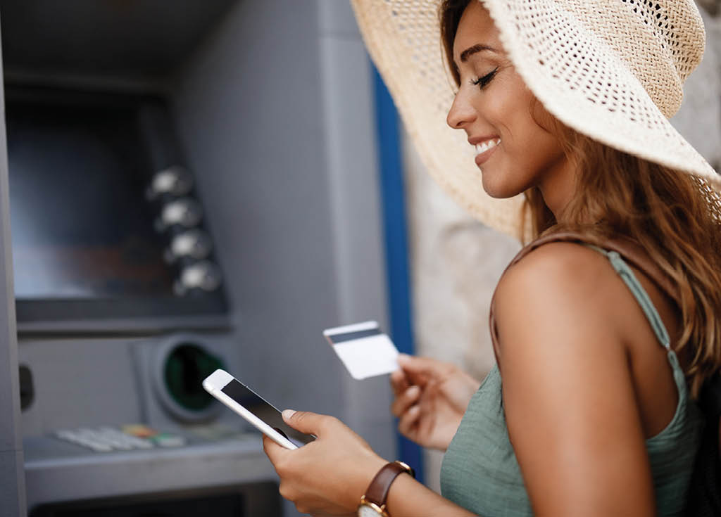 Smiling woman withdrawing money from cash machine while using smart phone 