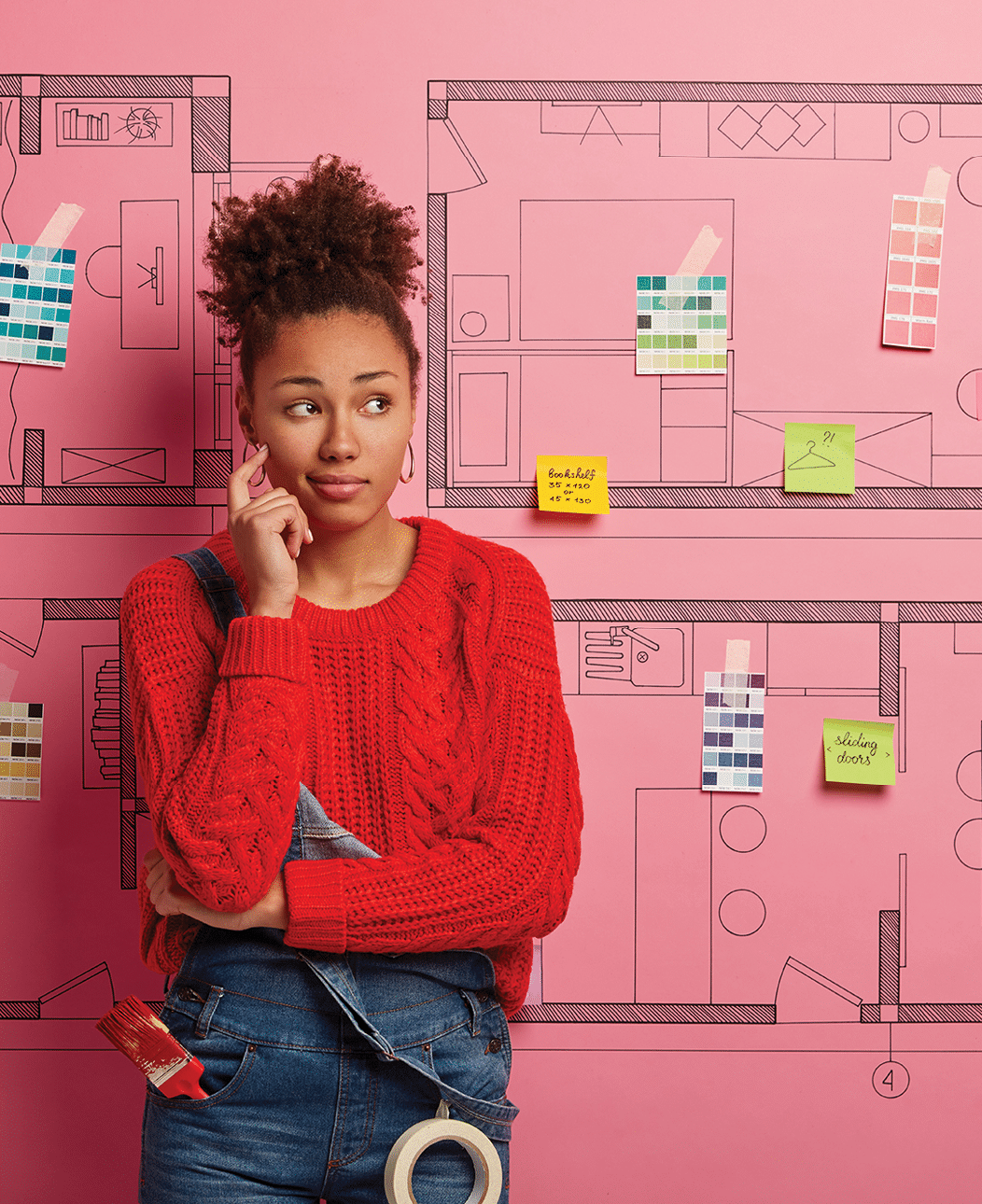 Unaware curly haired woman looks aside with thoughtful expression, has paint brush in pocket, moves in new house which needs improvement, thinks about buyig furniture, stands over pink background