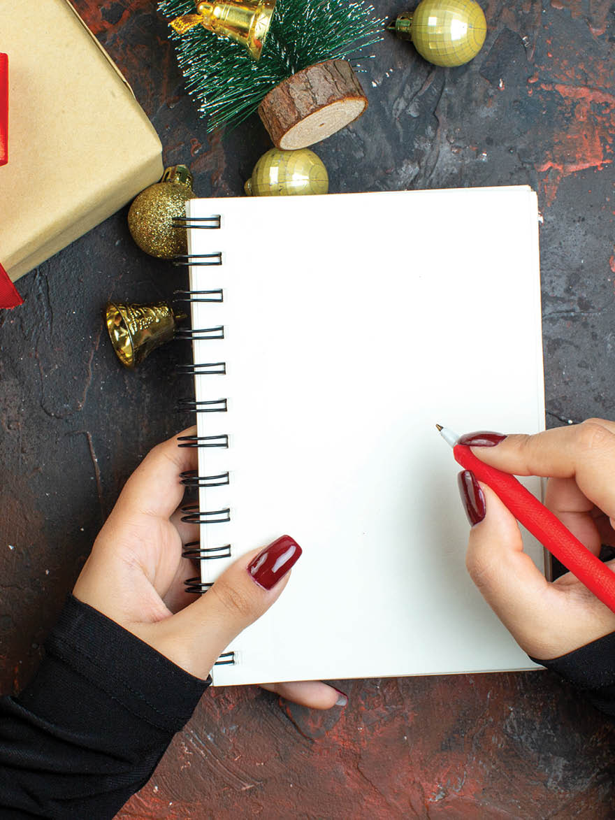 top view xmas gifts golden xmas balls mini xmas tree notebook and pen in woman hands on dark red table