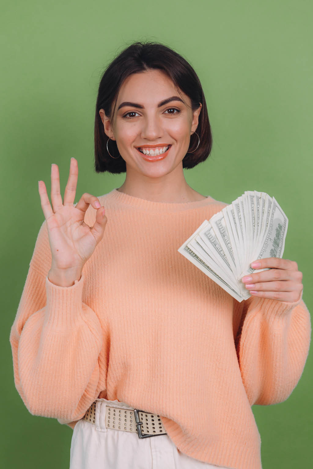 Young woman in casual peach sweater  isolated on green olive background  lucky holding fan of 100 dollar bills being excited to win cash prize showing OK sign