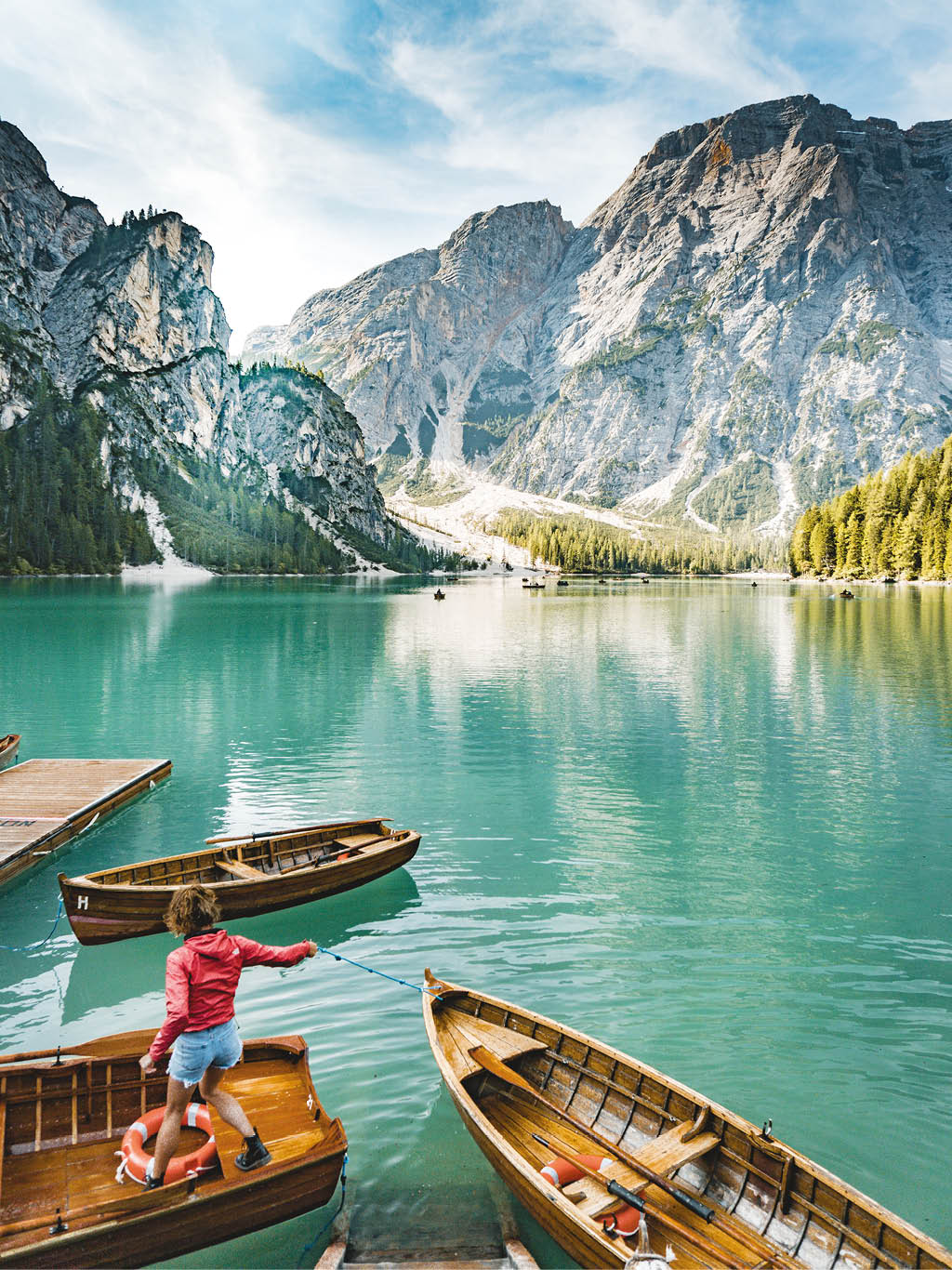 A beautiful shot of a lake with few boats with a female standing on one of them and amazing high rocky mountains in the background