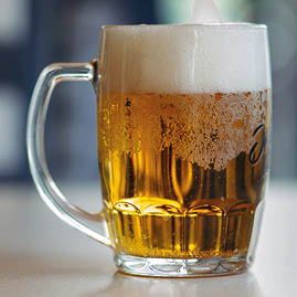 Horizontal photo of glass cup full of light fresh beer standing on smooth wooden surface. Cold summer drink for day heat. Fresh with thick high foam. Blurred background. Concept of drinks shooting.