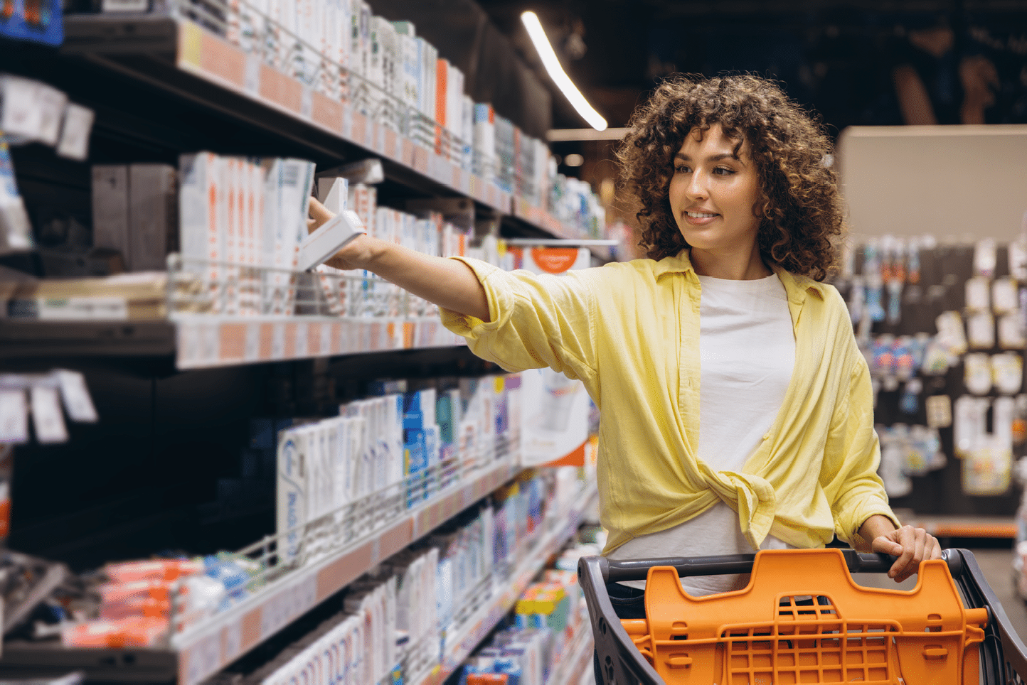 Young woman pushing shopping cart, reaching for toothpaste on shelf in aisle of large retail store, consumer buying