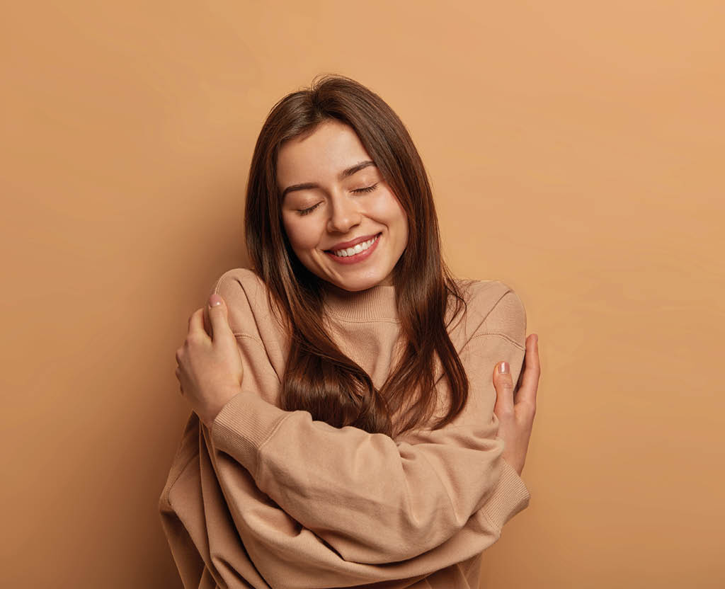 I love myself. Pretty smiling woman embraces herself gently, expresses self love, keeps eyes closed with pleasure, feels comfortable and fullfilled, being egoistic person, isolated on brown background