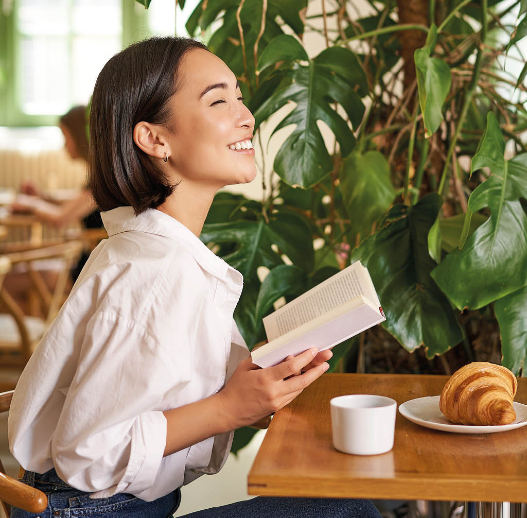 Vertical shot of asian brunette girl sitting elegant with book, reading in cafe and smiling, drinking coffee.