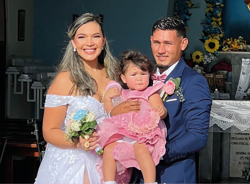 Una persona con vestido blanco de boda sostiene a un ni o peque o vestido de rosa mientras otra persona con traje azul los acompa a frente a un altar decorado con flores y una figura religiosa en el interior de un edificio. Descripci n generada con IA