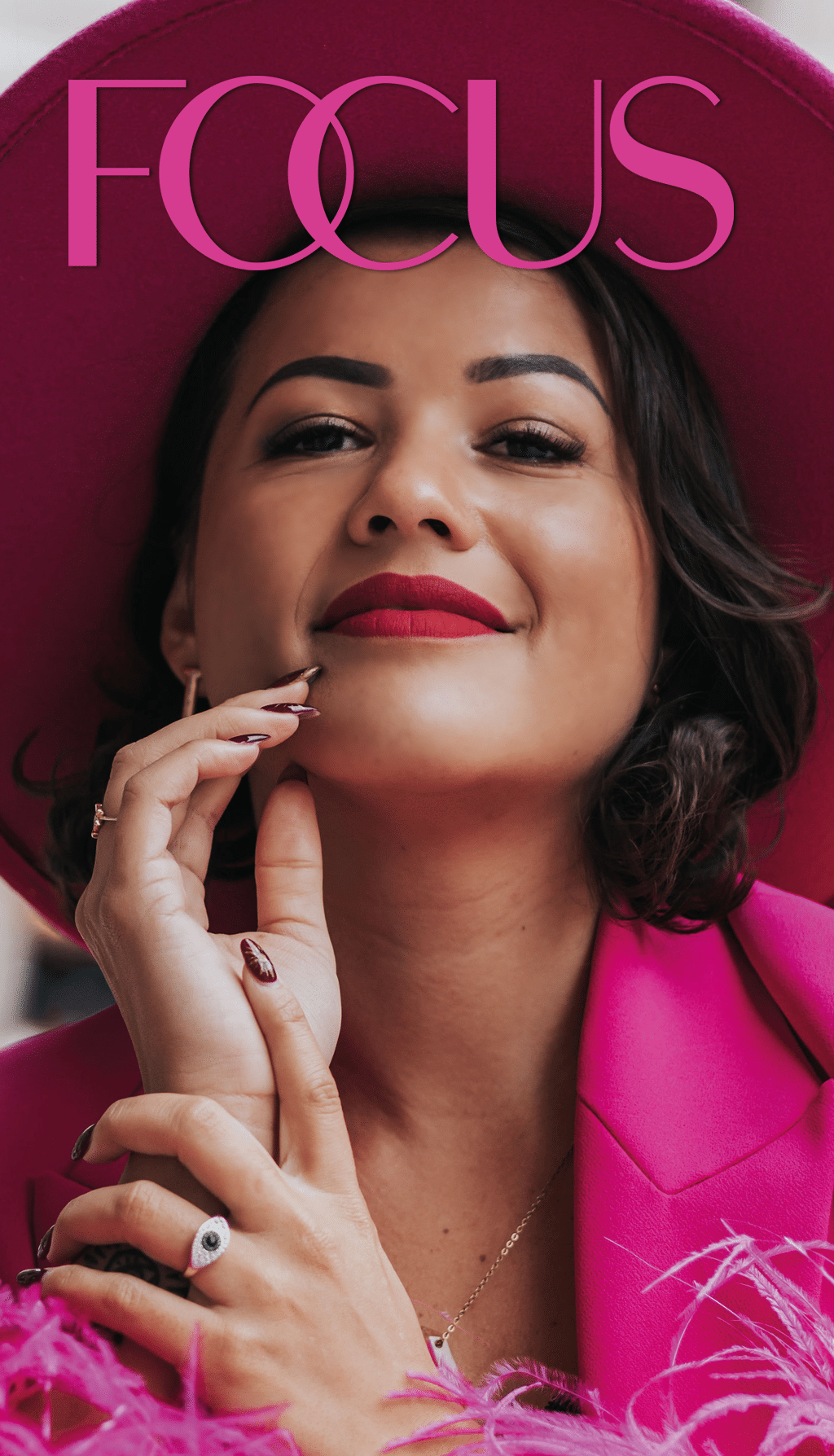 Persona con cabello corto y ondulado vistiendo un saco rosa brillante y un collar dorado mientras posa con las manos cerca del rostro frente a un fondo rojo con la palabra “FOCUS" en letras grandes. Descripci n generada con IA