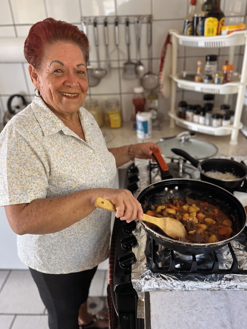Persona con cabello recogido y camiseta clara con estampado floral est cocinando guiso en una sart n grande sobre una estufa en una cocina dom stica.  Descripci n generada con IA