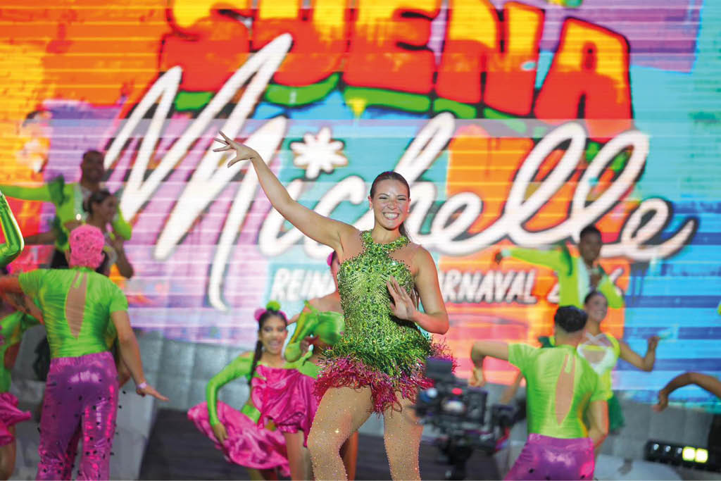Personas con trajes coloridos y brillantes bailan en un escenario con un fondo iluminado que muestra el texto “Sue a Michelle". Descripci n generada con IA