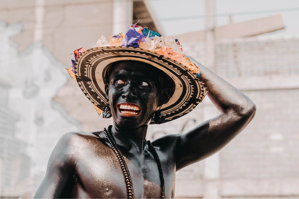 Persona con el torso desnudo y pintado de negro que lleva un sombrero tradicional decorado con flores de colores en un entorno urbano. Descripci n generada con IA