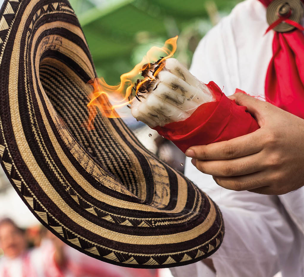 Una persona sostiene un sombrero tradicional decorado y una vela encendida envuelta en tela roja mientras viste una camisa blanca y un pa uelo rojo al cuello. Descripci n generada con IA