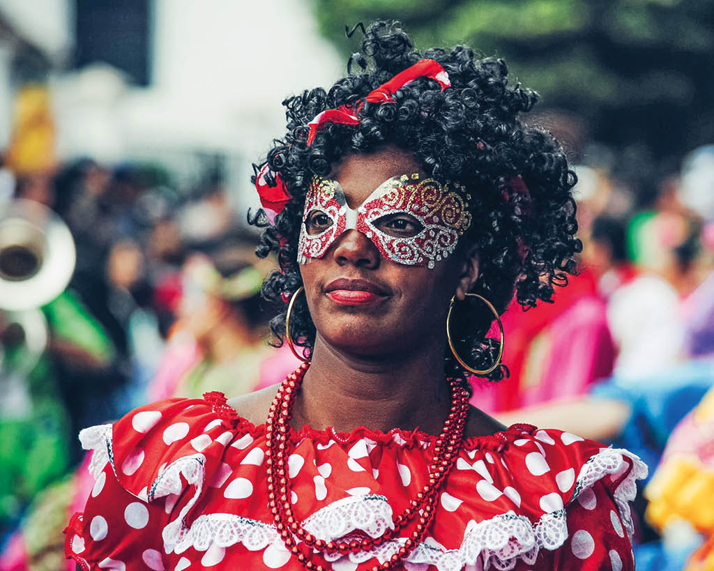 Persona con cabello rizado y vestido tradicional rojo con lunares blancos y encaje blanco, usando collar y accesorios, en un evento al aire libre con gente al fondo. Descripci n generada con IA