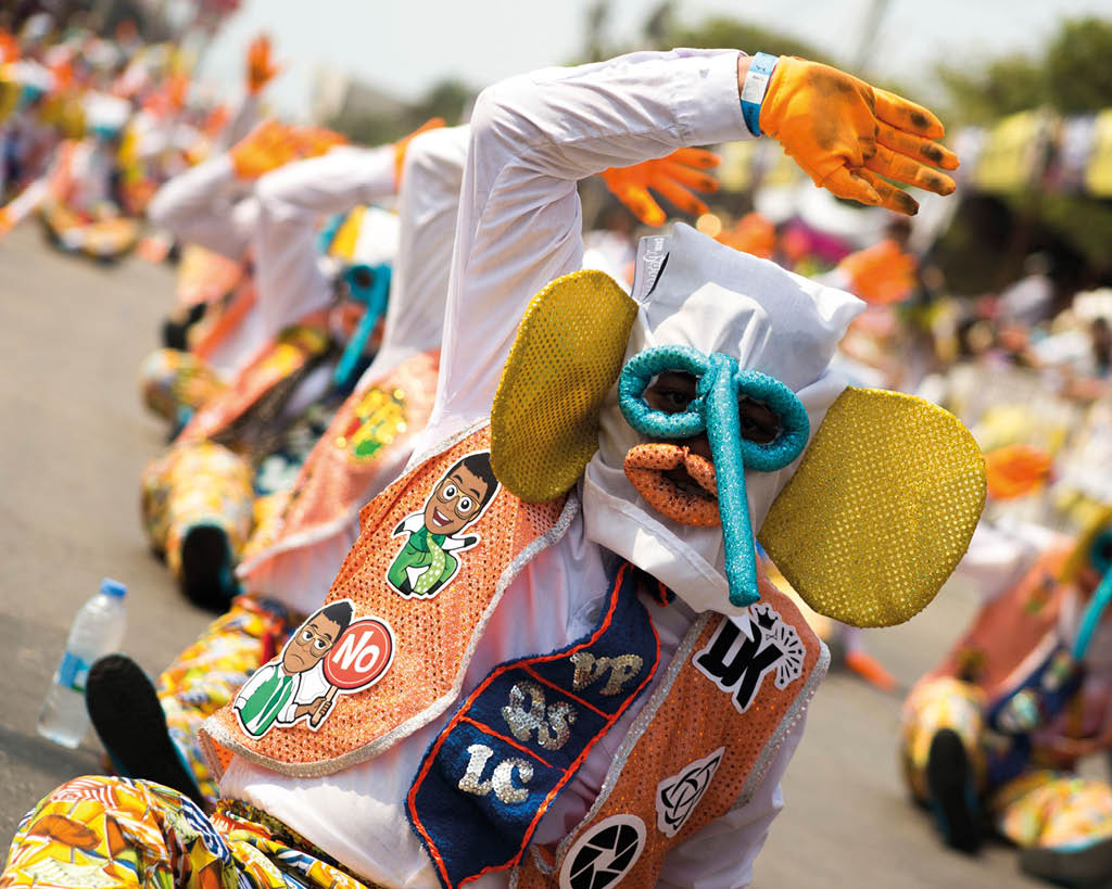 Personas vestidas con trajes coloridos y guantes naranjas participan en una danza tradicional al aire libre en una calle. Descripci n generada con IA