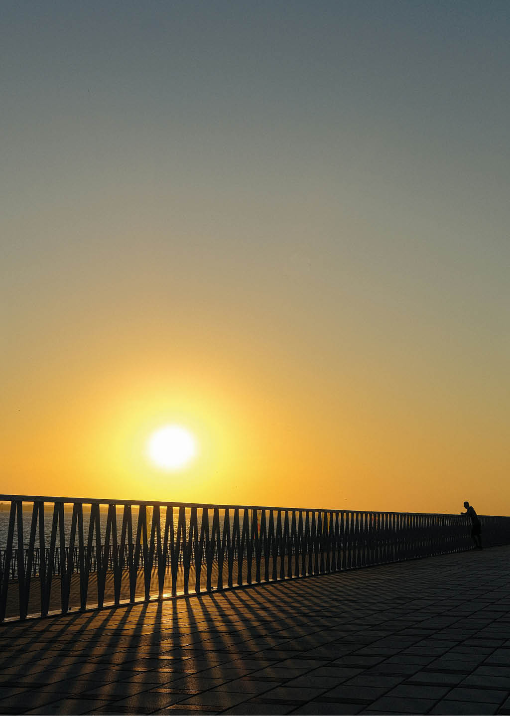 Un atardecer con el sol cerca del horizonte iluminando un camino adoquinado con una barandilla met lica y una persona apoyada en ella mirando hacia el paisaje. Descripci n generada con IA