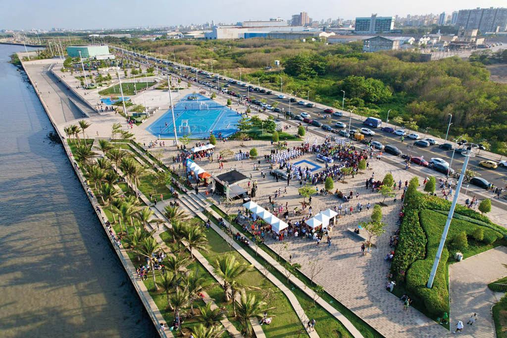 Un grupo de personas reunidas en un espacio p blico al aire libre con carpas blancas, reas verdes, palmeras y una cancha deportiva azul visible en la esquina superior izquierda.  Descripci n generada con IA