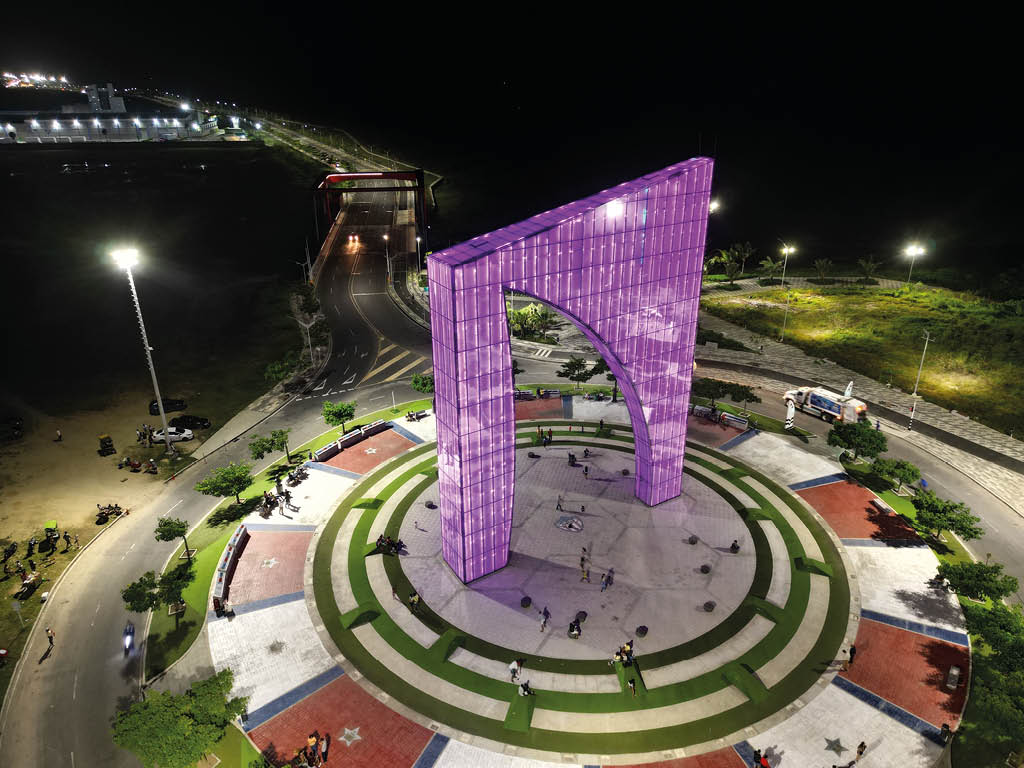 Monumento iluminado en color p rpura en una plaza circular con personas alrededor durante la noche y calles con vegetaci n y farolas encendidas. Descripci n generada con IA