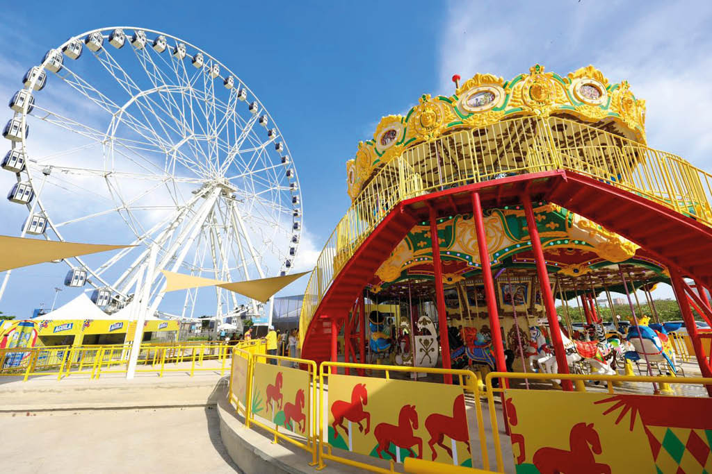 Una feria al aire libre con una noria blanca grande y un carrusel colorido decorado con caballos y detalles dorados bajo un cielo azul con algunas nubes. Descripci n generada con IA