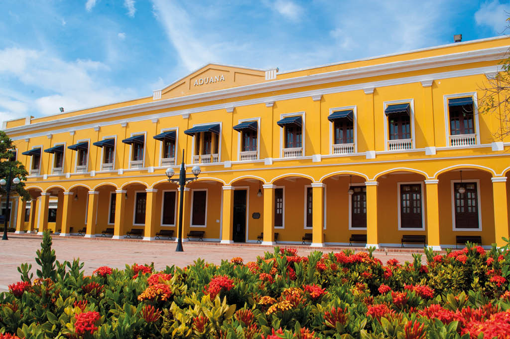 Edificio hist rico amarillo con arcos y ventanas con toldos azules frente a un jard n con flores rojas bajo un cielo azul con nubes. Descripci n generada con IA