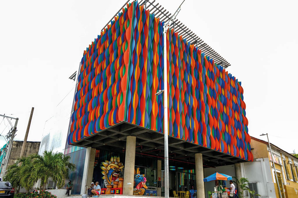 Edificio moderno con fachada decorada en patrones ondulados de colores rojo, azul y naranja, con personas sentadas y caminando en la entrada bajo un toldo azul y naranja. Descripci n generada con IA