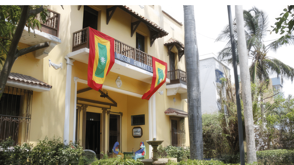 Dos personas con ropa tradicional est n sentadas frente a un edificio amarillo con dos banderas colgadas y un letrero que dice Museo Bhimayana. Descripci n generada con IA