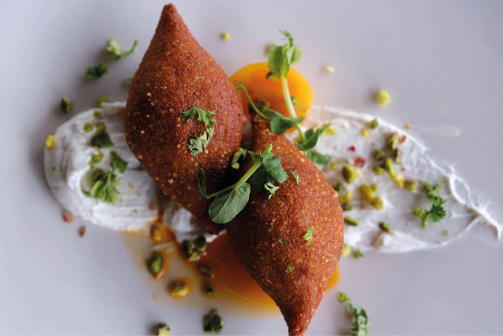 Dos croquetas fritas decoradas con hierbas frescas sobre una base de salsa blanca y trozos peque os de vegetales en un plato blanco. Descripci n generada con IA