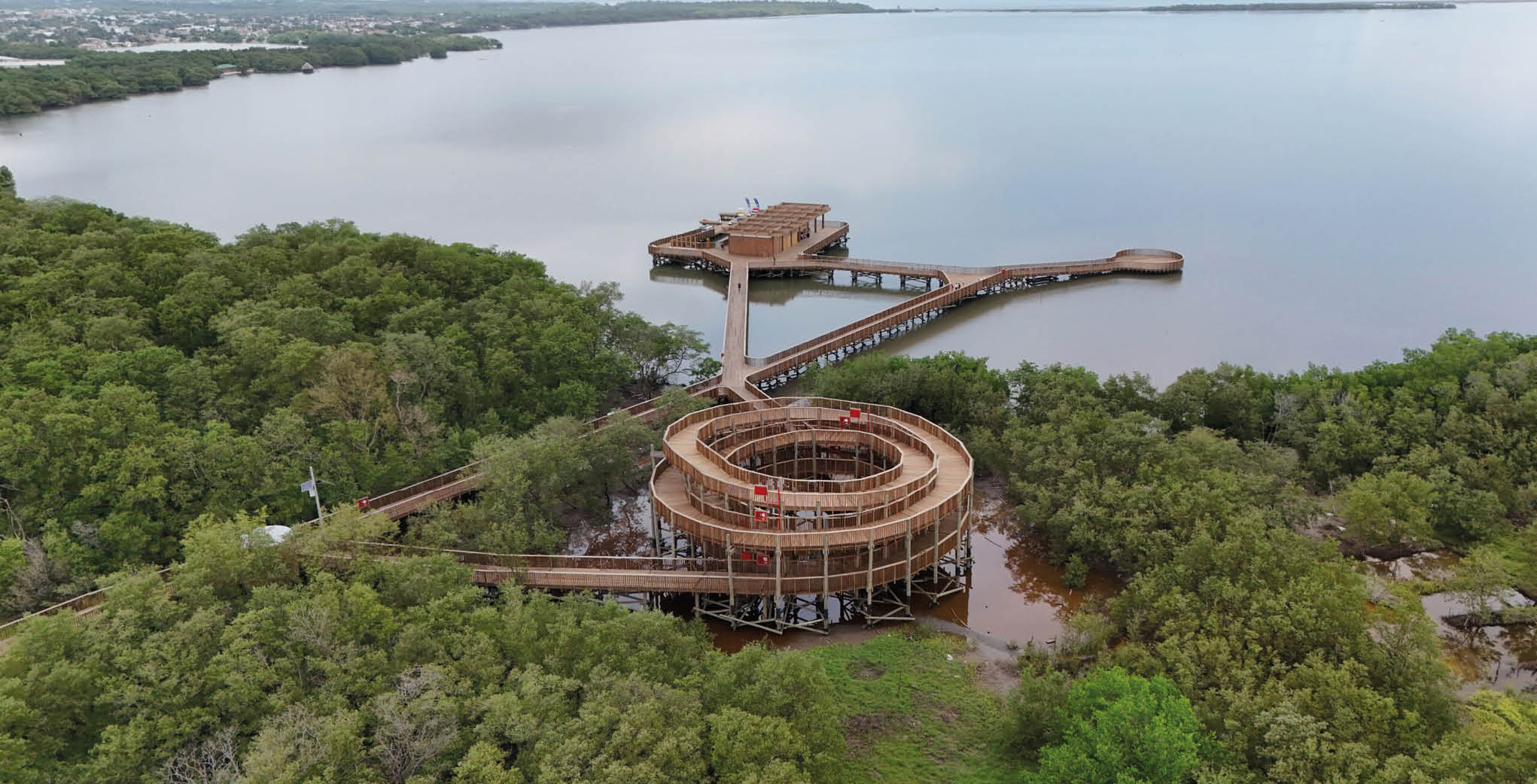Una estructura circular en construcci n rodeada de rboles junto a un muelle que se extiende sobre un cuerpo de agua tranquilo.  Descripci n generada con IA