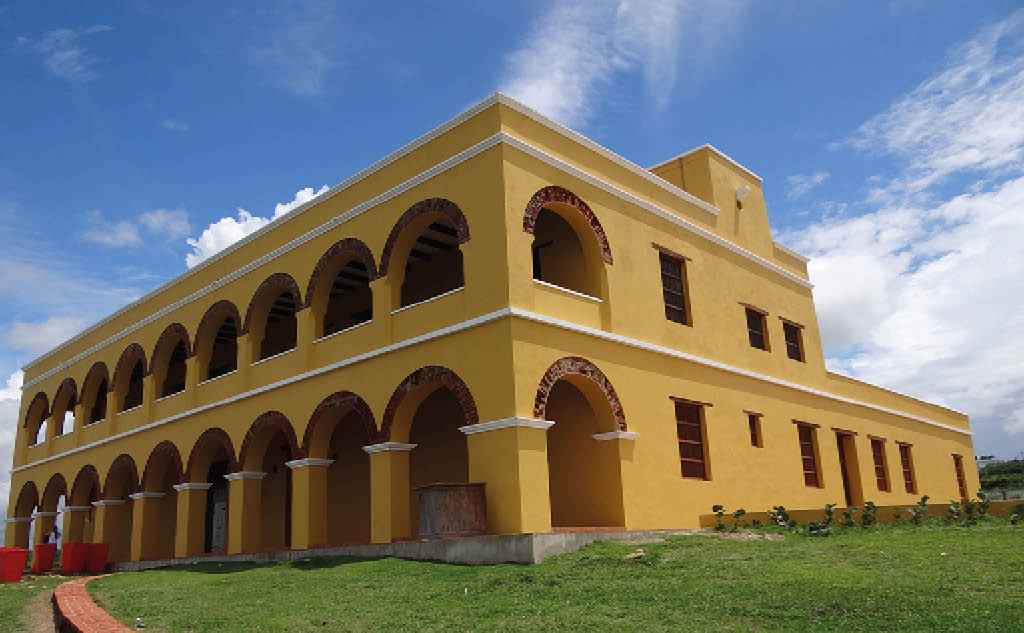 Edificio hist rico de dos pisos con arcos y paredes amarillas bajo un cielo azul con algunas nubes. Descripci n generada con IA