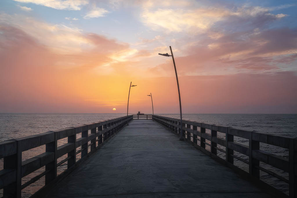 Un muelle de madera con farolas que se extiende hacia el mar bajo un cielo al atardecer con tonos anaranjados y nubes dispersas. Descripci n generada con IA