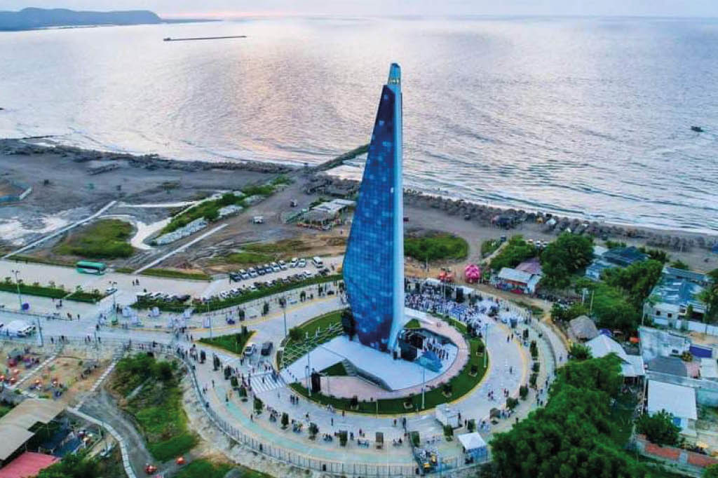 Un monumento alto y azul en forma de torre se encuentra en un rea circular rodeada de personas y veh culos cerca de la costa con el mar y un cielo nublado al fondo.  Descripci n generada con IA
