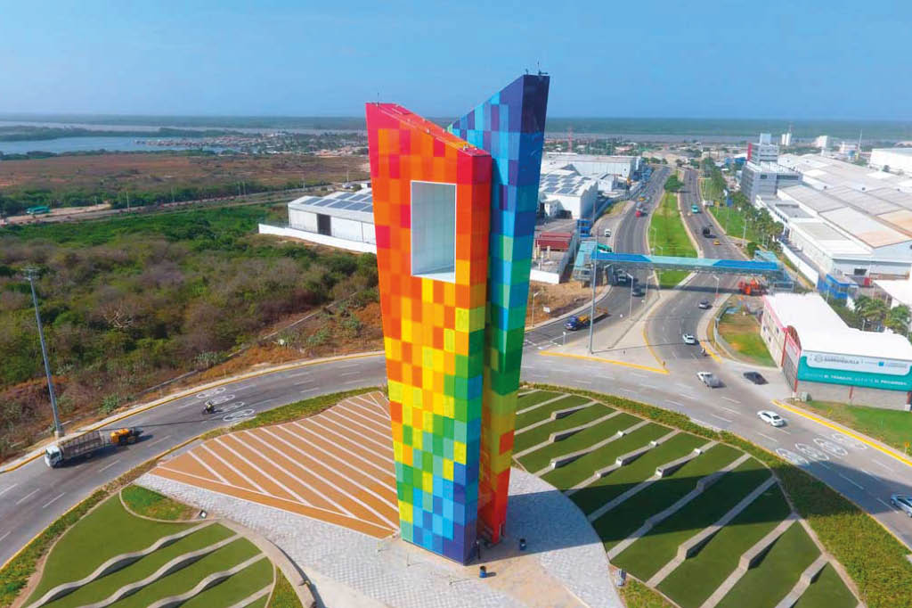 Una torre alta y delgada con un dise o de cuadros en colores brillantes est situada en una rotonda rodeada de carreteras y  reas verdes en un entorno urbano.  Descripci n generada con IA