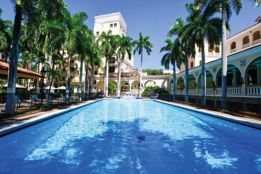 Piscina al aire libre rodeada de palmeras y edificios con arquitectura colonial bajo un cielo despejado. Descripci n generada con IA