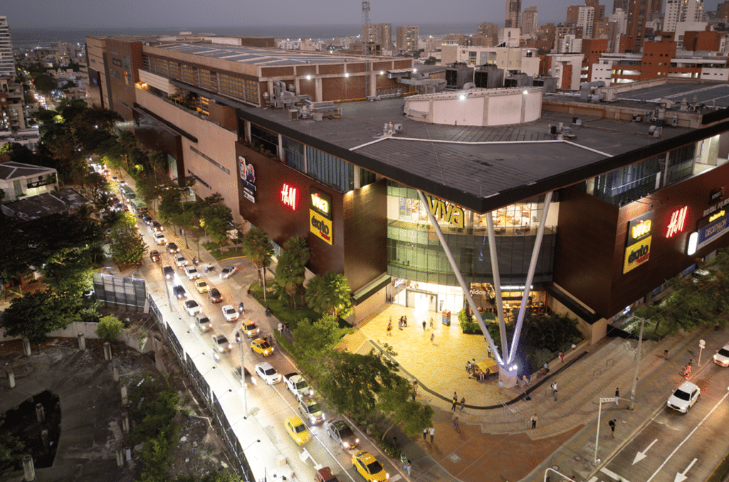 Centro comercial moderno iluminado al anochecer con tr fico de veh culos y personas caminando en las aceras. Descripci n generada con IA