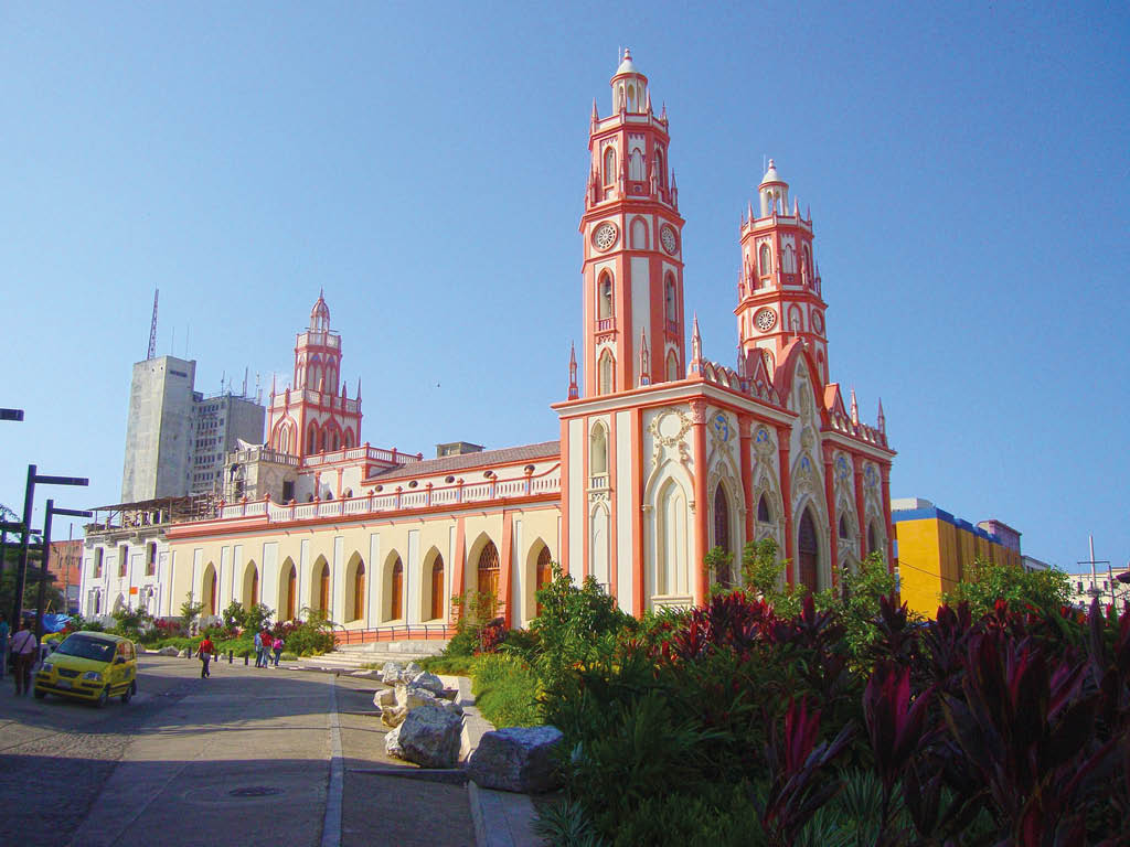 Edificio hist rico de estilo neog tico con torres altas y detalles en color rojo y blanco junto a una calle con personas y vegetaci n bajo un cielo despejado. Descripci n generada con IA