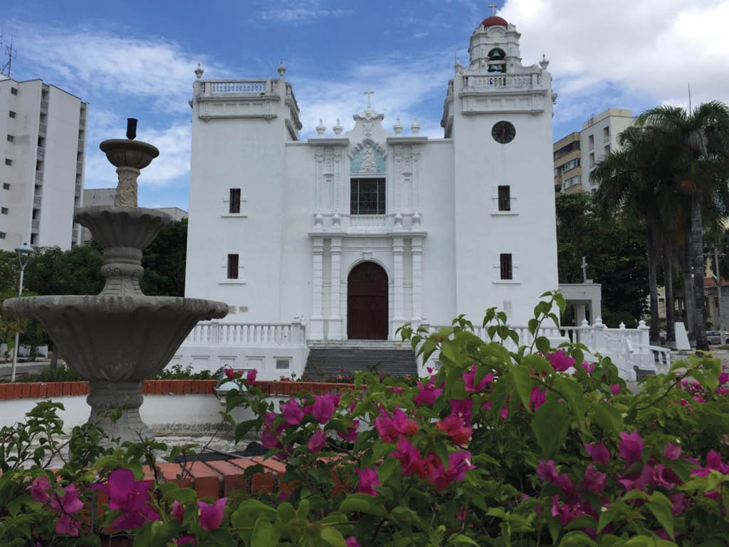 Una iglesia blanca de estilo colonial con una fuente de piedra y flores moradas en primer plano rodeada de edificios y palmeras bajo un cielo parcialmente nublado. Descripci n generada con IA