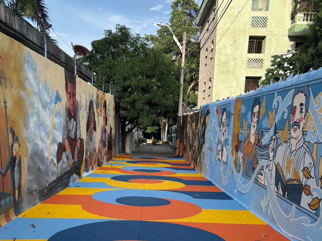 Un pasillo al aire libre con murales coloridos que representan personas y un suelo pintado con c rculos y franjas en tonos azul, naranja y amarillo. Descripci n generada con IA