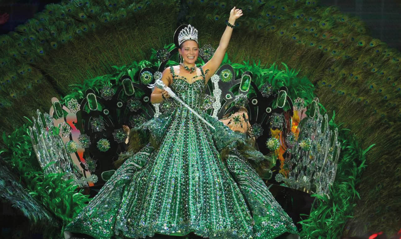 Persona con vestido verde brillante y corona sentada en una estructura decorativa que simula un gran abanico o flor en un evento festivo. Descripci n generada con IA