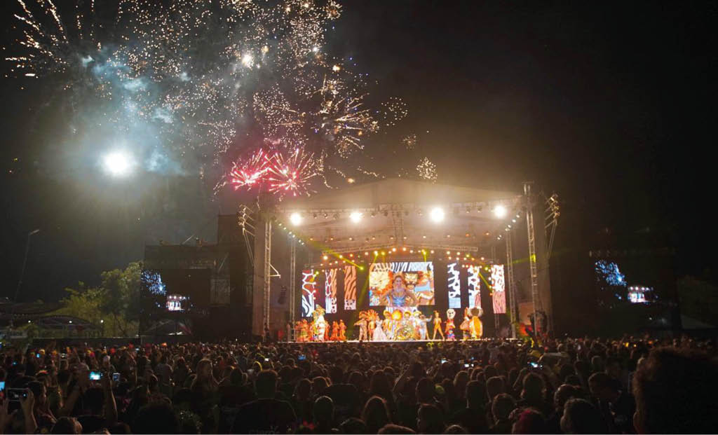 Un grupo de personas en un escenario iluminado con pantallas LED y fuegos artificiales explotan en el cielo nocturno mientras una multitud observa. Descripci n generada con IA