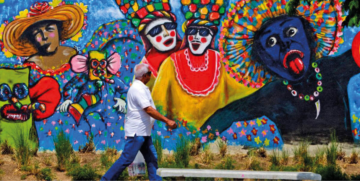 Una persona con ropa casual camina frente a un mural colorido que muestra figuras festivas con m scaras y trajes tradicionales en un entorno al aire libre. Descripci n generada con IA