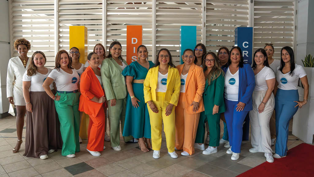Un grupo de personas vestidas con trajes de colores brillantes posan juntas en un espacio interior con paneles y carteles coloridos al fondo. Descripci n generada con IA