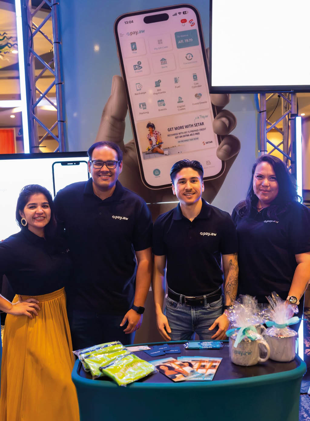 Cuatro personas sonrientes con camisetas negras posan detr s de una mesa con folletos y productos promocionales frente a un gran cartel que muestra un tel fono m vil con una aplicaci n abierta. Descripci n generada con IA