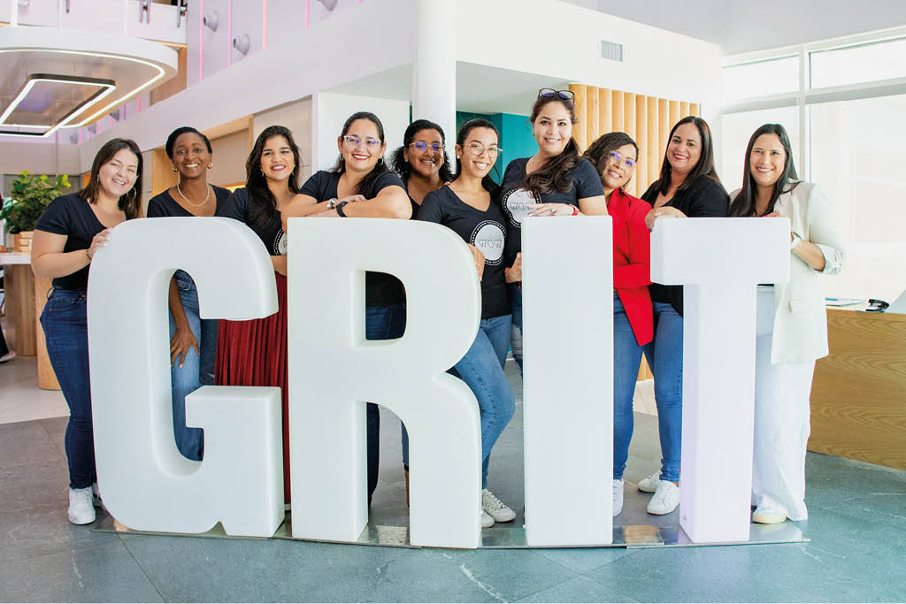 Un grupo de personas sonrientes posan juntas detr s de letras grandes que forman la palabra GRIT en un espacio interior moderno y bien iluminado. Descripci n generada con IA